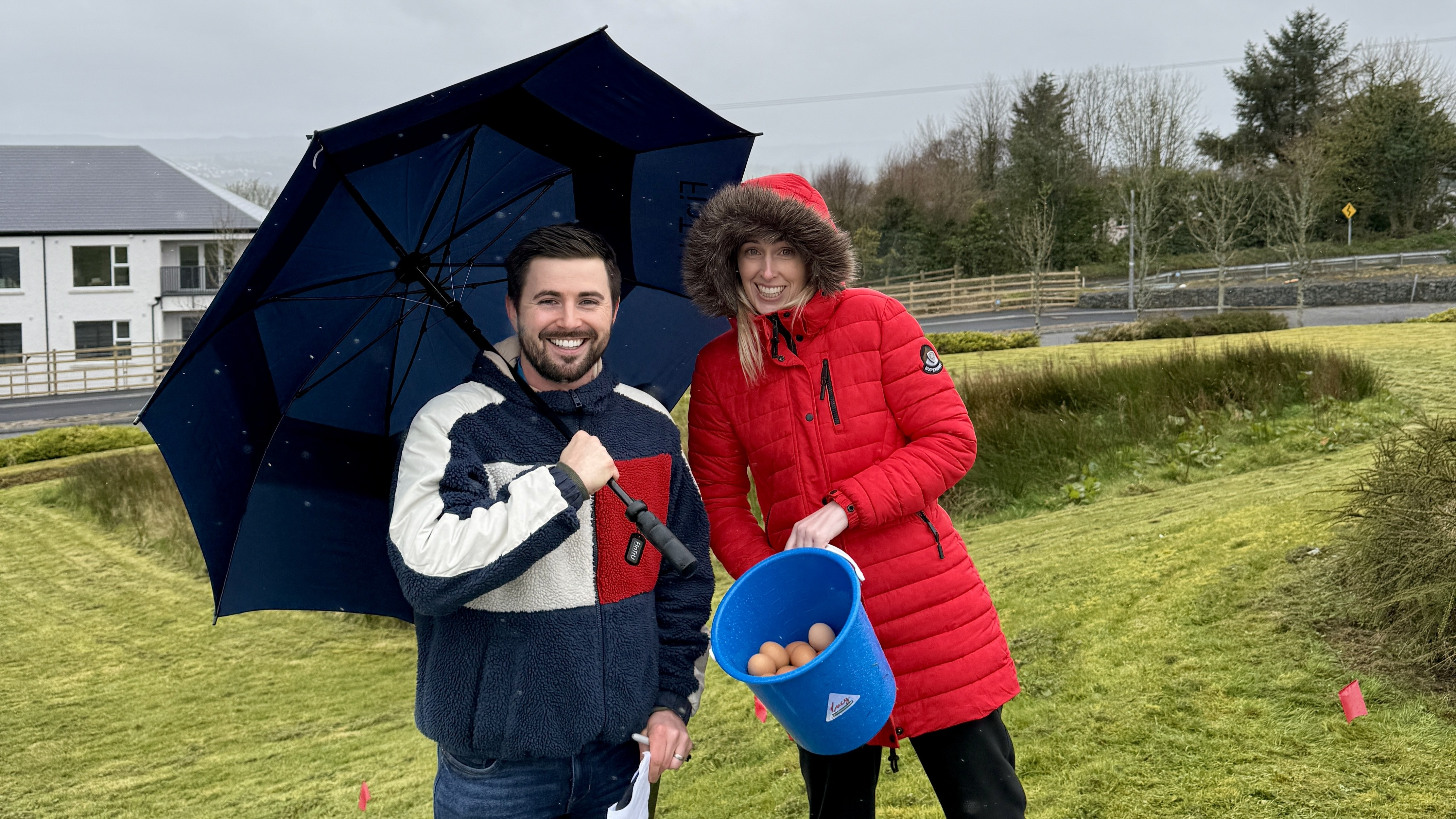 two people under an umbrella holding a bucket of eggs on a grass embankment
