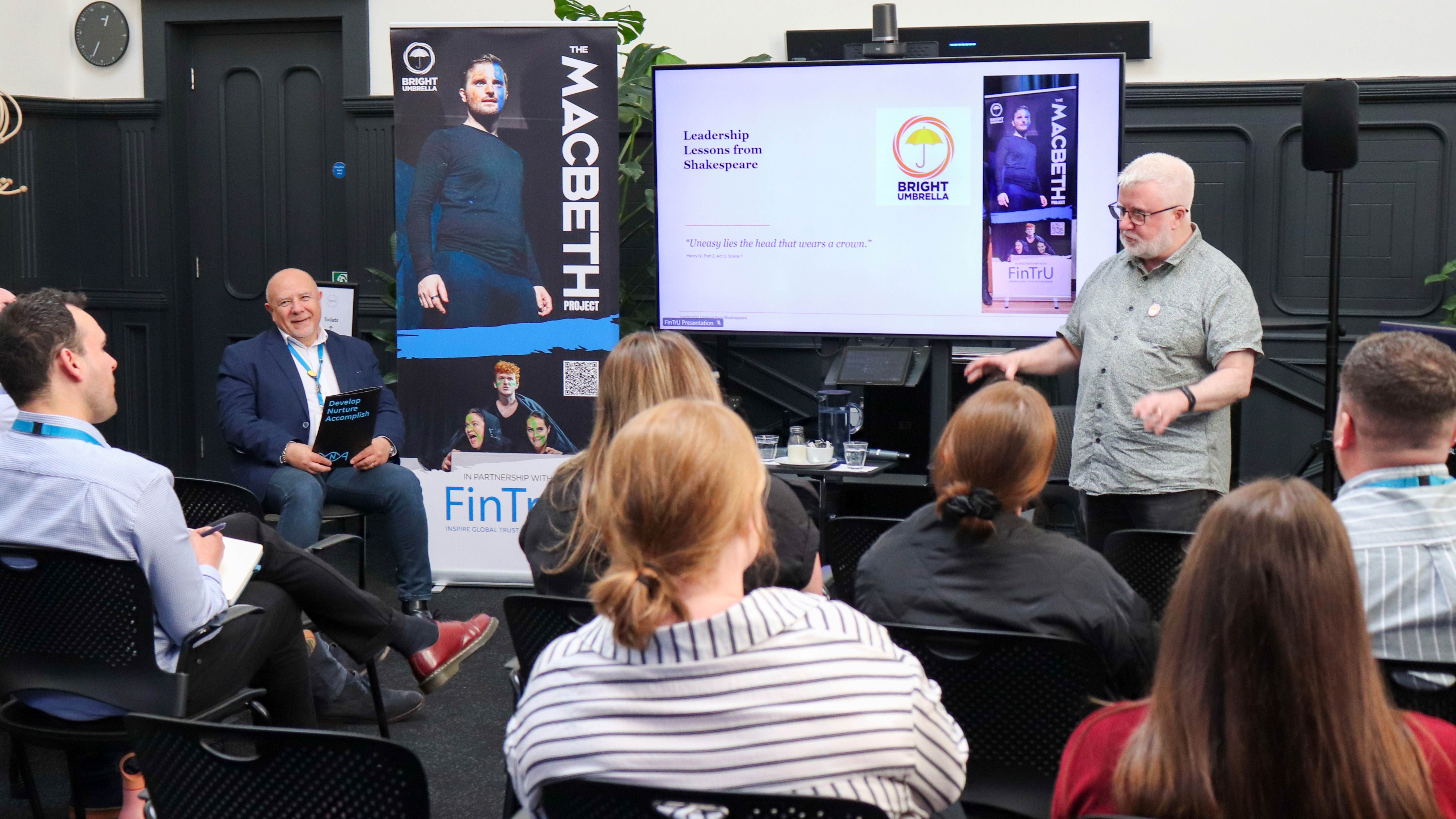 a man standing in front of a room full of seated people with a pull down banner of Macbeth and screen behind him